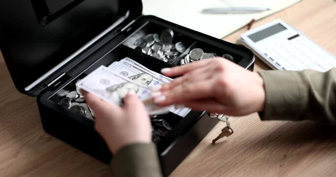 Woman takes dollar bills out of cash register of accountant with safety lock and key. Black box with banknotes and coins on wooden table slow motion