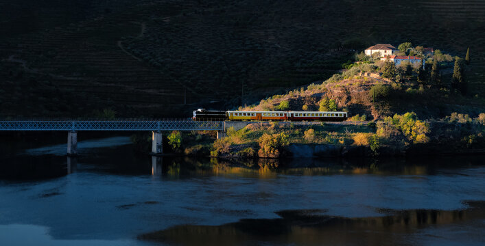 Historic touristic train in the douro valley, passing on the iron bridge and illuminated by the rays of the early morning sun - Port wine vineyards in the background mountains - Pocinho, Portugal