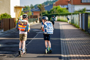 Two school kid boys in safety helmet riding with scooter in the city with backpack on sunny day. Happy children in colorful clothes biking on way to school. Safe way for kids outdoors to school