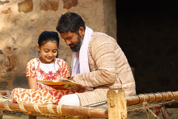 Happy rural Indian father and daughter sitting together on a cot outside their cottage/home/house. Father is teaching his daughter how to read with a book in his hand