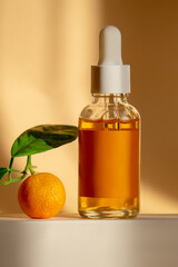 Moisturizing orange serum for face with vitamin C in glass bottle with dropper lid on white square stand. Unmarked cosmetic container next to an orange. Vertical frame, close-up.