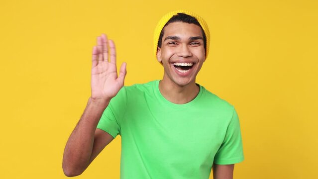 Young man of African American ethnicity wear green t-shirt hat look around for friend find wave meet greet with hand as notices someone say hello hi isolated on plain yellow background studio portrait