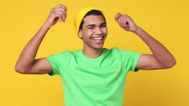 Young man of African American ethnicity wear green t-shirt hat dance waving expressive gesticulating hand fool around have fun enjoy isolated on plain yellow background studio People lifestyle concept