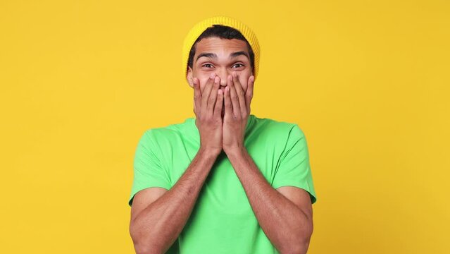 Young man of African American ethnicity wear green t-shirt hat look camera surprised ask what wow omg no way do winner gesture say yes celebrating put hands on face isolated on plain yellow background