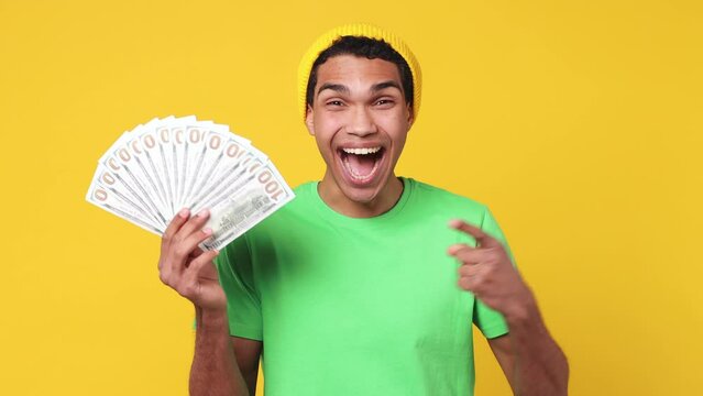 Happy young man of African American ethnicity wearing green t-shirt hat hold fan of cash money in dollar banknotes doing winner gesture celebrate isolated on plain yellow background studio portrait