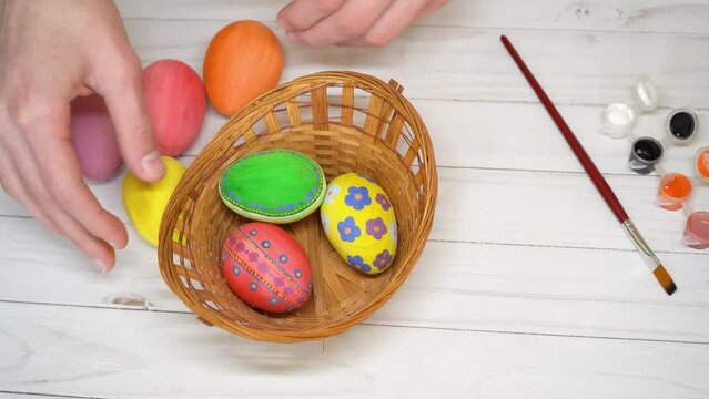 Human hands take out of basket colored eggs on white table background, top view. Happy Easter concept. Unrecognized caucasian man preparing beautiful holy symbols for religious holiday