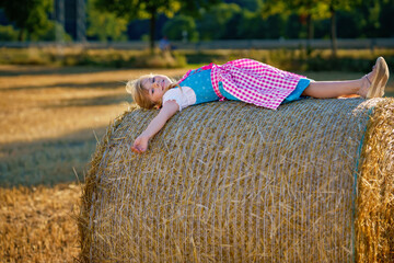 Cute little kid girl in traditional Bavarian costume in wheat field. Happy child with hay bale...