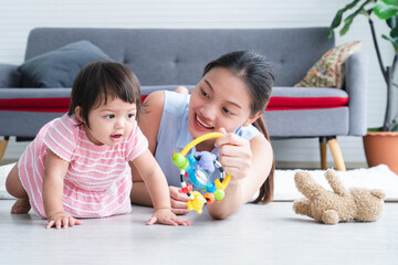 Asian young mother and Caucasian 7 months baby newborn girl playing with development toy and bear doll. Single mom lying on floor smiling playing with little daughter, crawling in living room at home