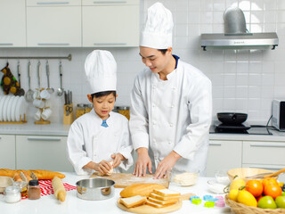 Asian young little boy chef in uniform with white tall cook hat standing learning preparing massaging flour dough on wooden cutting board while male cooking teacher helping teaching in home kitchen