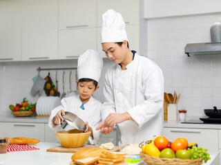 Asian young little boy pastry chef in white uniform with tall cook hat standing using stainless filter preparing sifting flour on wooden board while male cooking teacher helping teaching in kitchen