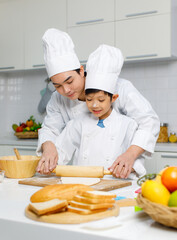 Asian young little boy chef in uniform with white tall cook hat standing learning preparing massaging flour dough on wooden cutting board while male cooking teacher helping teaching in home kitchen