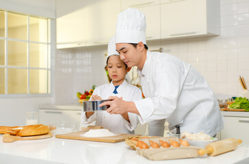 Asian young little boy pastry chef in white uniform with tall cook hat standing using stainless filter preparing sifting flour on wooden board while male cooking teacher helping teaching in kitchen