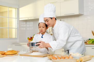 Asian young little boy pastry chef in white uniform with tall cook hat standing using stainless filter preparing sifting flour on wooden board while male cooking teacher helping teaching in kitchen