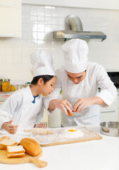 Asian young little boy pastry chef in white uniform with tall cook hat standing learning preparing egg into sifting flour on wooden board while male cooking teacher smiling help teaching in kitchen