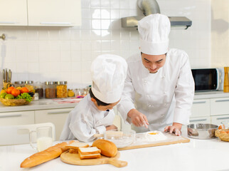 Asian young little boy pastry chef in white uniform with tall cook hat standing learning preparing...