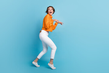 Fototapeta na wymiar Full size photo of pretty young girl look around sneaking tiptoes play dinosaur wear trendy orange outfit isolated on blue color background
