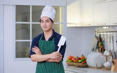 Millennial Asian male pastry chef wearing white tall cook hat and apron standing smiling posing in home white kitchen full of fruits vegetables and bread on cooking counter.