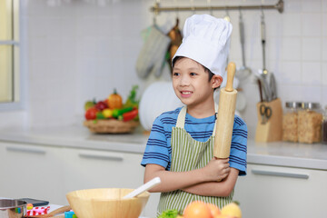 Millennial Asian little boy chef wearing tall white cook hat and apron standing posing in home white kitchen full of fruits vegetables and bread on cooking counter.