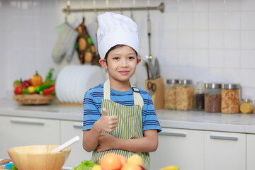 Millennial Asian little boy chef wearing tall white cook hat and apron standing posing in home white kitchen full of fruits vegetables and bread on cooking counter.