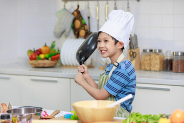 Millennial Asian young little boy chef wearing white tall cook hat and apron standing smiling holding cooking pan posing taking photo while preparing cooking fresh meal at counter in home kitchen