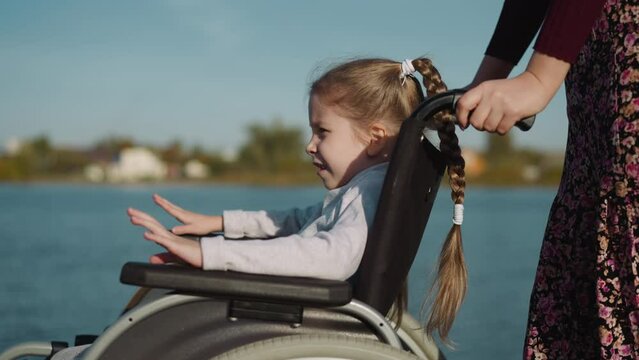 Mother and daughter with paralysis walk near pond. Blonde girl claps hands on armrests of wheelchair looking into distance on blurred background closeup
