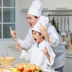 Asian young little boy chef and male cooking teacher in uniform with white tall cook hat standing holding rock star hands sign up open mouth wide screaming shouting posing together in home kitchen