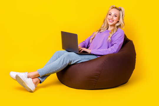 Full length photo of cheerful positive woman wear violet shirt sitting bean bad typing emails device isolated yellow color background