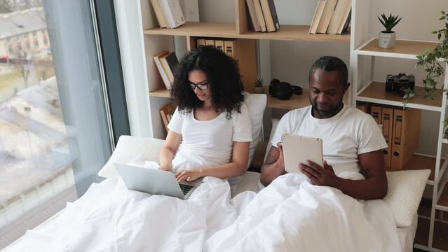 African american romantic partners using electronic devices while lounging on wide bed near panoramic window. Beautiful woman with laptop and relaxed man with tablet enjoying leisure time at home.