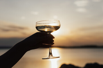 Woman's hand holding glass of wine with sea and beautiful sunset at background, Close-up. Summer evening relaxed mood.