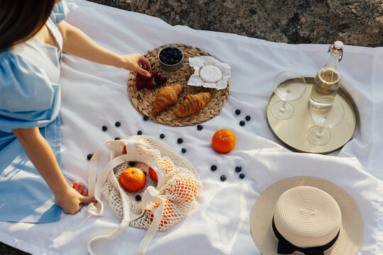 Cropped picture of a woman sitting on a picnic at nature. Wine and two wineglasses on the golden tray, eco shopping bag with oranges, straw hat and croissants, berries and cheese brie.