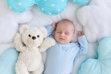 baby boy sleeping on the bed lying on his back with a soft teddy bear among the pillows in blue pajamas, healthy newborn sleep