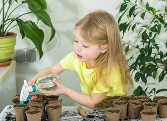 little child girl in yellow shirt watering plants in pots with soil from spray gun in light room....