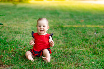 a laughing baby in summer on the green grass in a red bodysuit in the setting sun rejoices, space for text