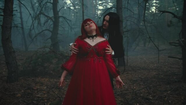 Night dark misty forest. gothic couple male vampire prince hugs fantasy woman princess by shoulders looks at her neck wants to bite taste blood. Lady girl medieval red dress. Man count in black tuxedo