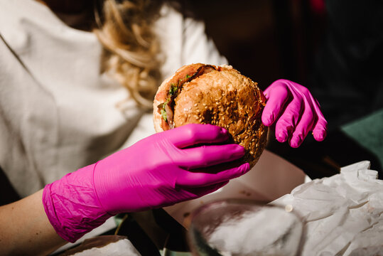 Girl eating and holding a burger in pink gloves, food photo, cheeseburger closeup. Burger party. Stylish birthday party in fast food restaurant. Woman eat cheeseburgers. Female enjoys tasty hamburger.