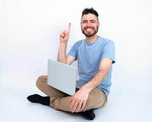 Young caucasian man with laptop sitting over white studio showing and pointing up with finger number one while smiling confident and happy.