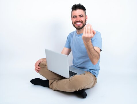 Young caucasian man with laptop sitting over white studio inviting to come with hand. Happy that you came
