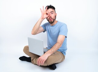 Young caucasian man with laptop sitting over white studio doing ok gesture shocked with surprised face, eye looking through fingers. Unbelieving expression.