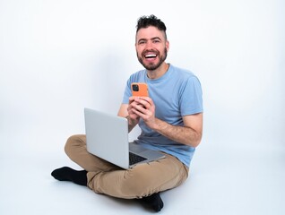Excited Young caucasian man with laptop sitting over white studio holding smartphone and looking amazed to the camera after receiving good news.