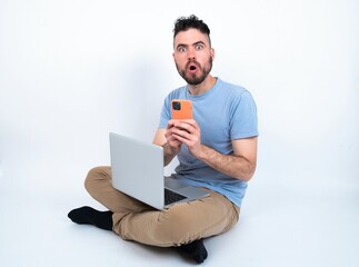 Shocked Young caucasian man with laptop sitting over white studio opens mouth hold phone reading advert unbelievable big shopping prices