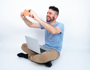 Young caucasian man with laptop sitting over white studio taking a selfie to post it on social...