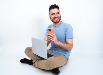 Young caucasian man with laptop sitting over white studio hold telephone hands read good youth news look empty space advert