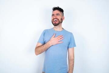 Fototapeta premium Joyful Young caucasian man wearing blue T-shirt over white background expresses positive emotions recalls something funny keeps hand on chest and giggles happily.