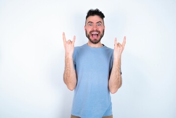 Young caucasian man wearing blue T-shirt over white background makes rock n roll sign looks self confident and cheerful enjoys cool music at party. Body language concept.