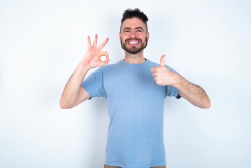 Young caucasian man wearing blue T-shirt over white background feeling happy, amazed, satisfied and surprised, showing okay and thumbs up gestures, smiling