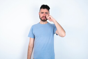 Disappointed dejected Young caucasian man wearing blue T-shirt over white background wipes tears stands stressed with gloomy expression. Negative emotion