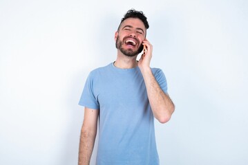 Funny Young caucasian man wearing blue T-shirt over white background laughs happily, has phone conversation, being amused by friend, closes eyes.