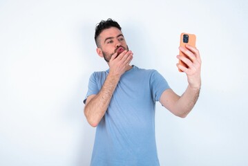 Young caucasian man wearing blue T-shirt over white background blows air kiss at camera of smartphone and takes selfie, sends mwah via online call.