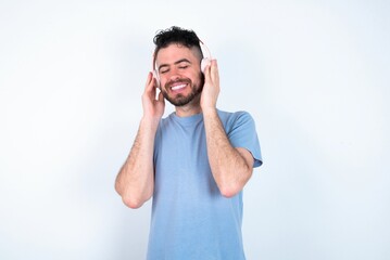 Pleased Young caucasian man wearing blue T-shirt over white background enjoys listening pleasant melody keeps hands on stereo headphones closes eyes. Spending free time with music