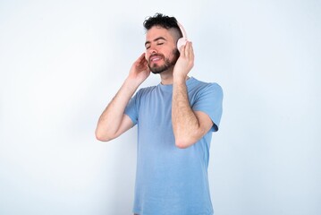 Fototapeta premium Young caucasian man wearing blue T-shirt over white background with headphones on her head, listens to music, enjoying favourite song with closed eyes, holding hands on headset.